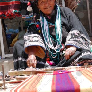 Indigenous woman with hat and necklaces working on loom smiles 1